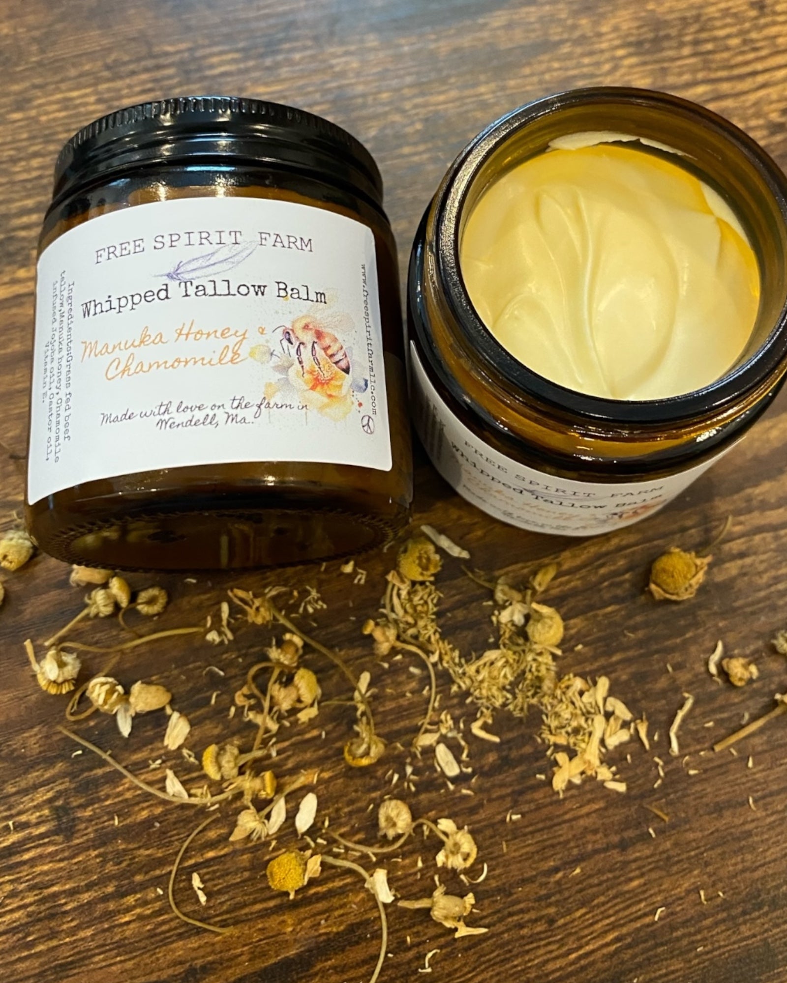 A jar of Whipped Tallow Balm with Manuka Honey & Chamomile on a wooden surface, with ingredients like chamomile flowers scattered around.