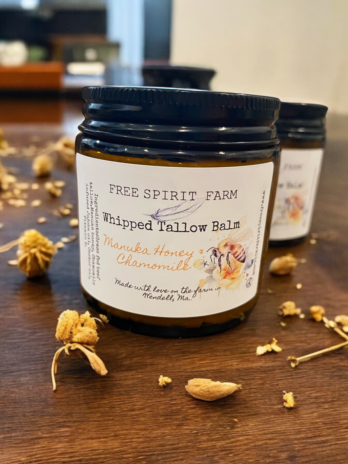 A jar of Whipped Tallow Balm with Manuka Honey & Chamomile on a wooden surface, with ingredients like chamomile flowers scattered around.