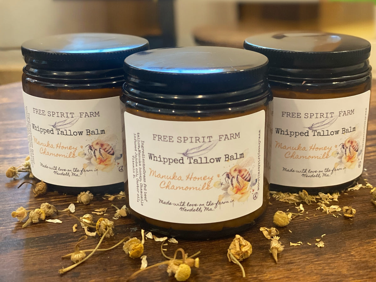 A jar of Whipped Tallow Balm with Manuka Honey & Chamomile on a wooden surface, with ingredients like chamomile flowers scattered around.