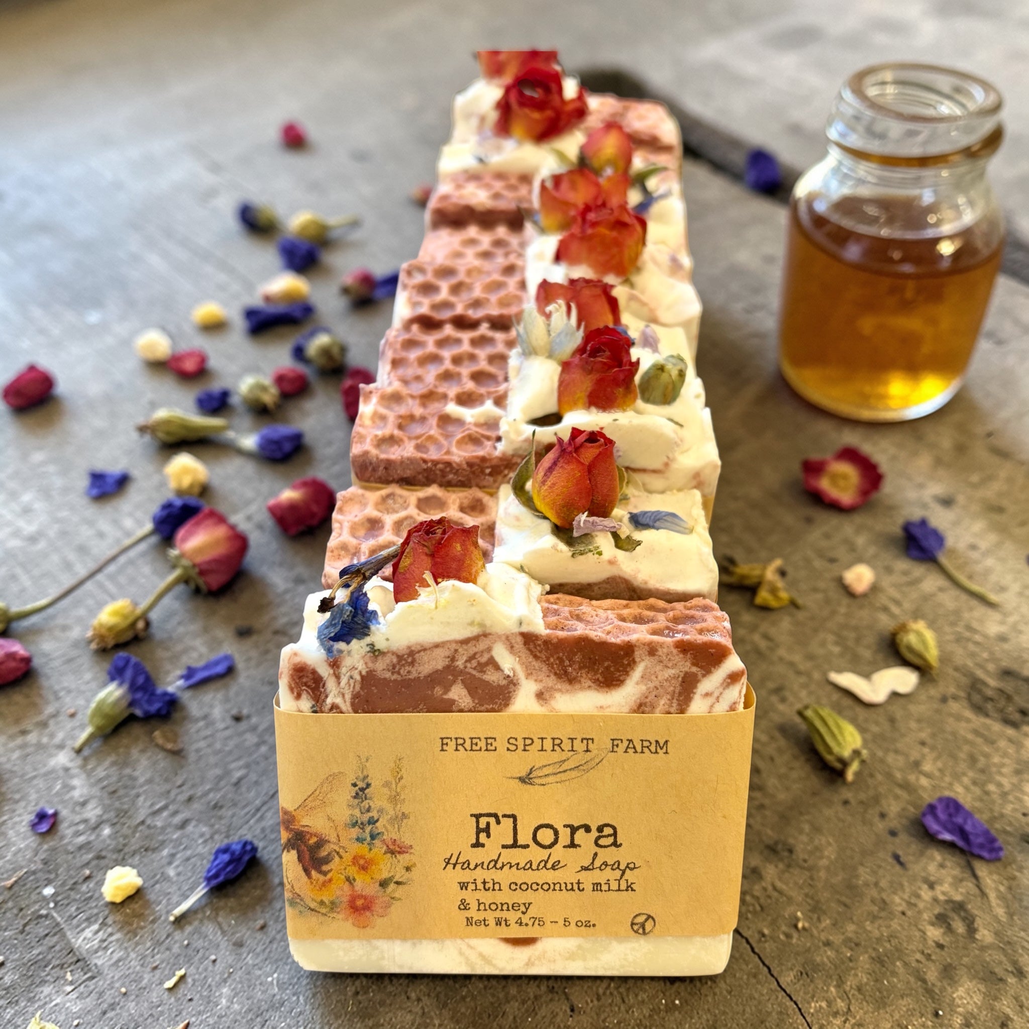 Row of handmade soaps with floral decorations on a rustic surface, accompanied by a jar of honey.