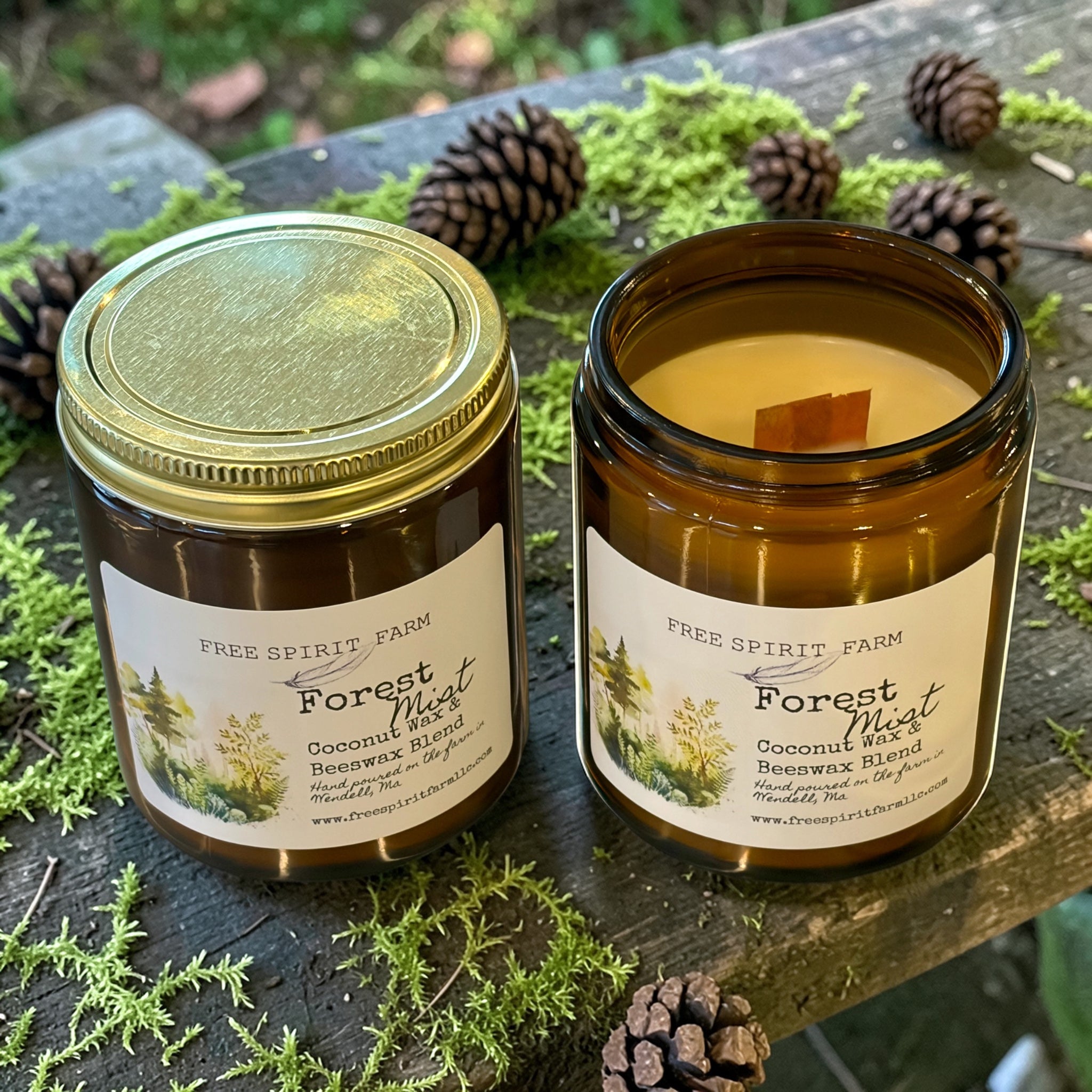 Two Free Spirit Farm candles, one in a jar and one in a glass container, on a wooden surface with pinecones and greenery.