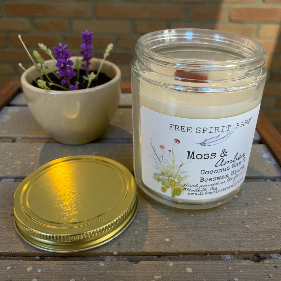 Candle jar with a label on a wooden surface next to a potted plant.