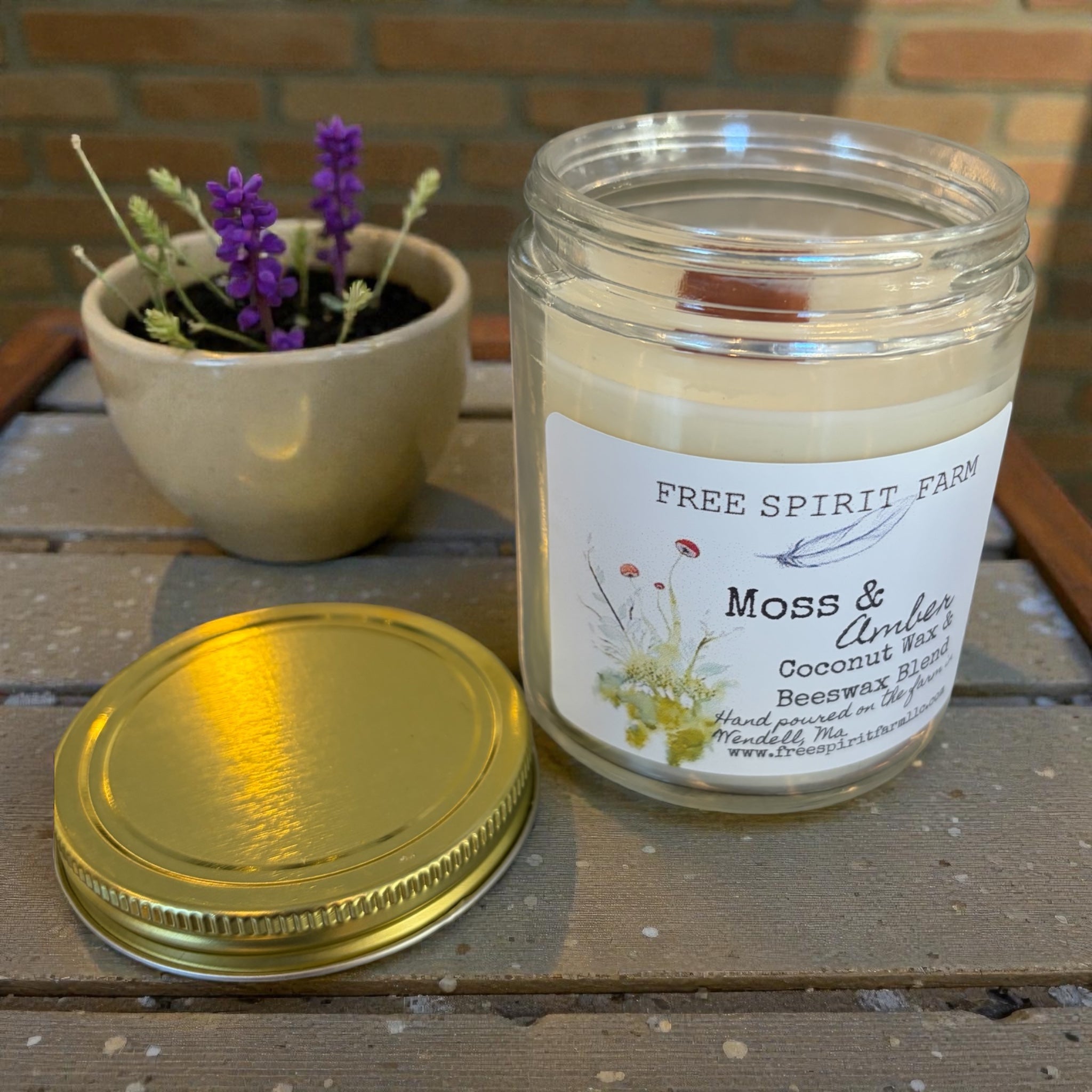Candle jar with a label on a wooden surface next to a potted plant.