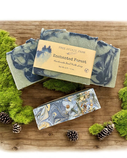 Handmade goat milk soap bar with green and blue swirls, topped with seeds and a touch of gold mica, placed on a wooden surface with moss and pine cones around.