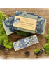 Handmade goat milk soap bar with green and blue swirls, topped with seeds and a touch of gold mica, placed on a wooden surface with moss and pine cones around.