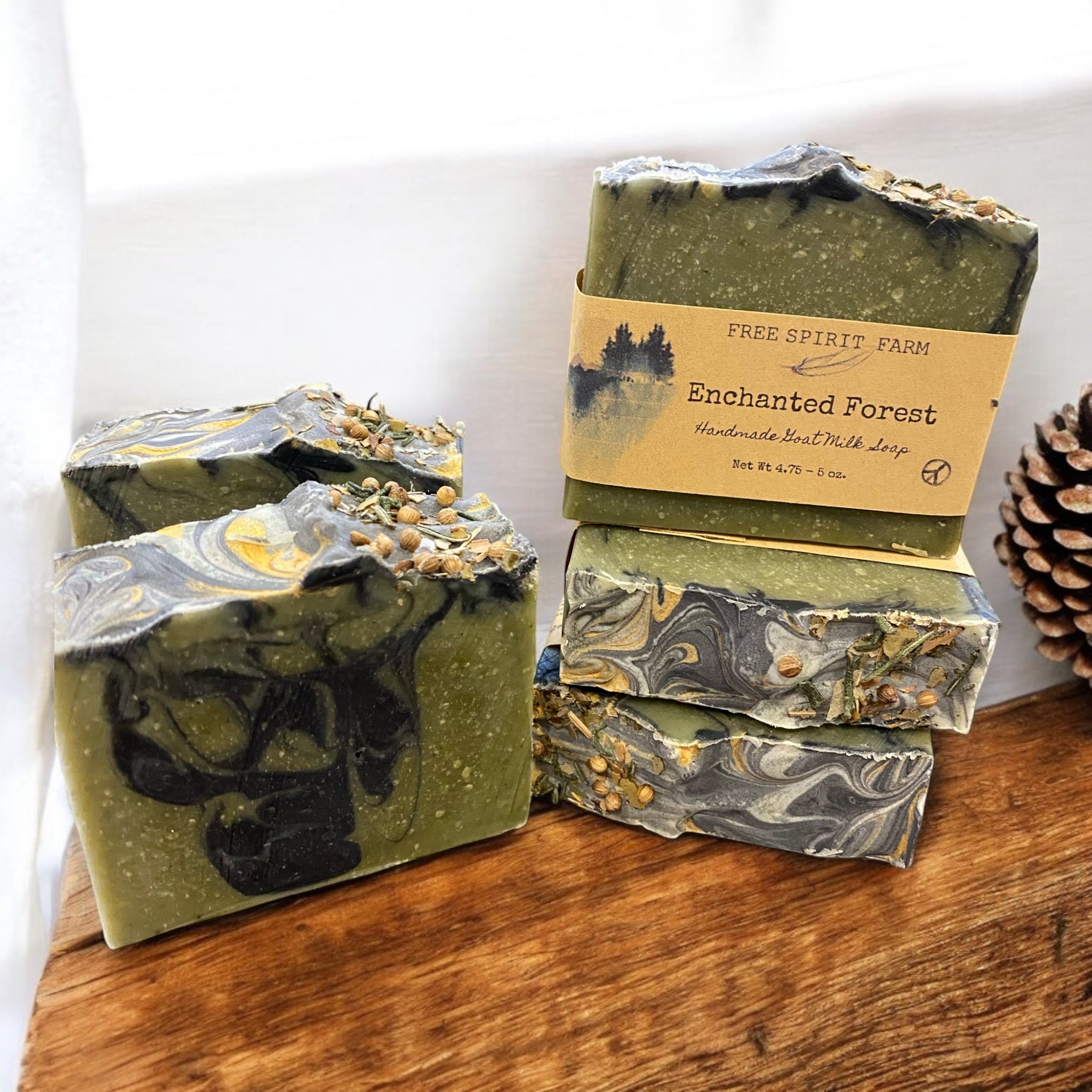 Handmade goat milk soap bar with green and blue swirls, topped with seeds and a touch of gold mica, placed on a wooden surface with moss and pine cones around.