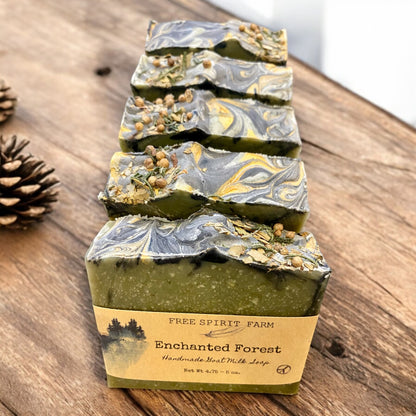 Handmade goat milk soap bar with green and blue swirls, topped with seeds and a touch of gold mica, placed on a wooden surface with moss and pine cones around.
