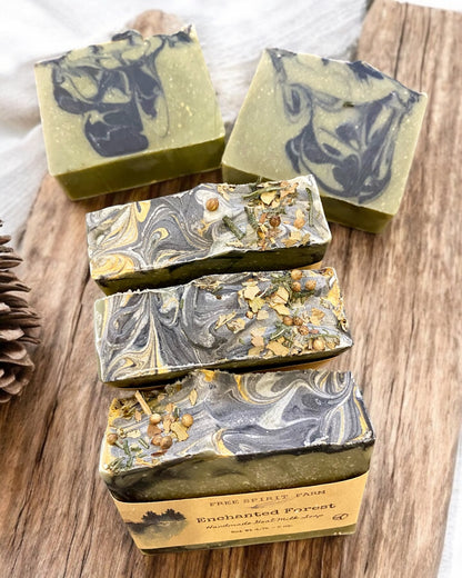 Handmade goat milk soap bar with green and blue swirls, topped with seeds and a touch of gold mica, placed on a wooden surface with moss and pine cones around.