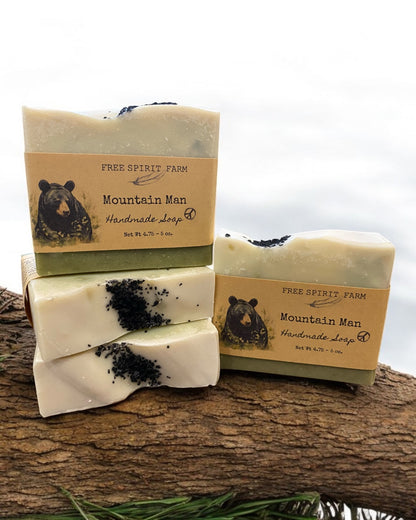 A bar of handmade vegan soap with a mountain man theme, featuring a beige color and black accents resembling soil or dirt, placed on a wooden surface with packaging in the background.