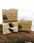A bar of handmade vegan soap with a mountain man theme, featuring a beige color and black accents resembling soil or dirt, placed on a wooden surface with packaging in the background.