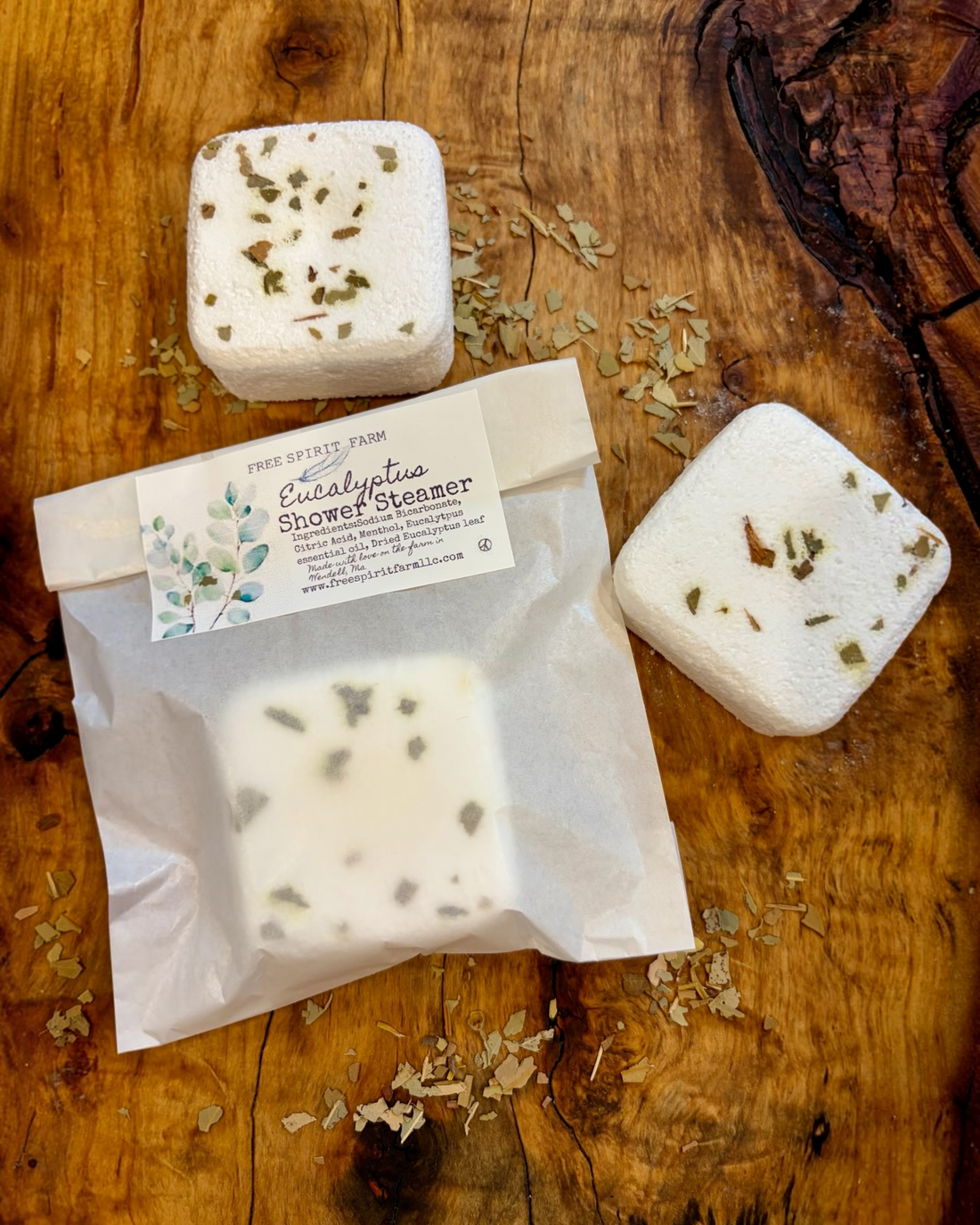 Free Spirit Farm  square white shower steamer bars with dried botanical inclusions, one shown unpackaged and one inside a clear resealable pouch with a white label reading &