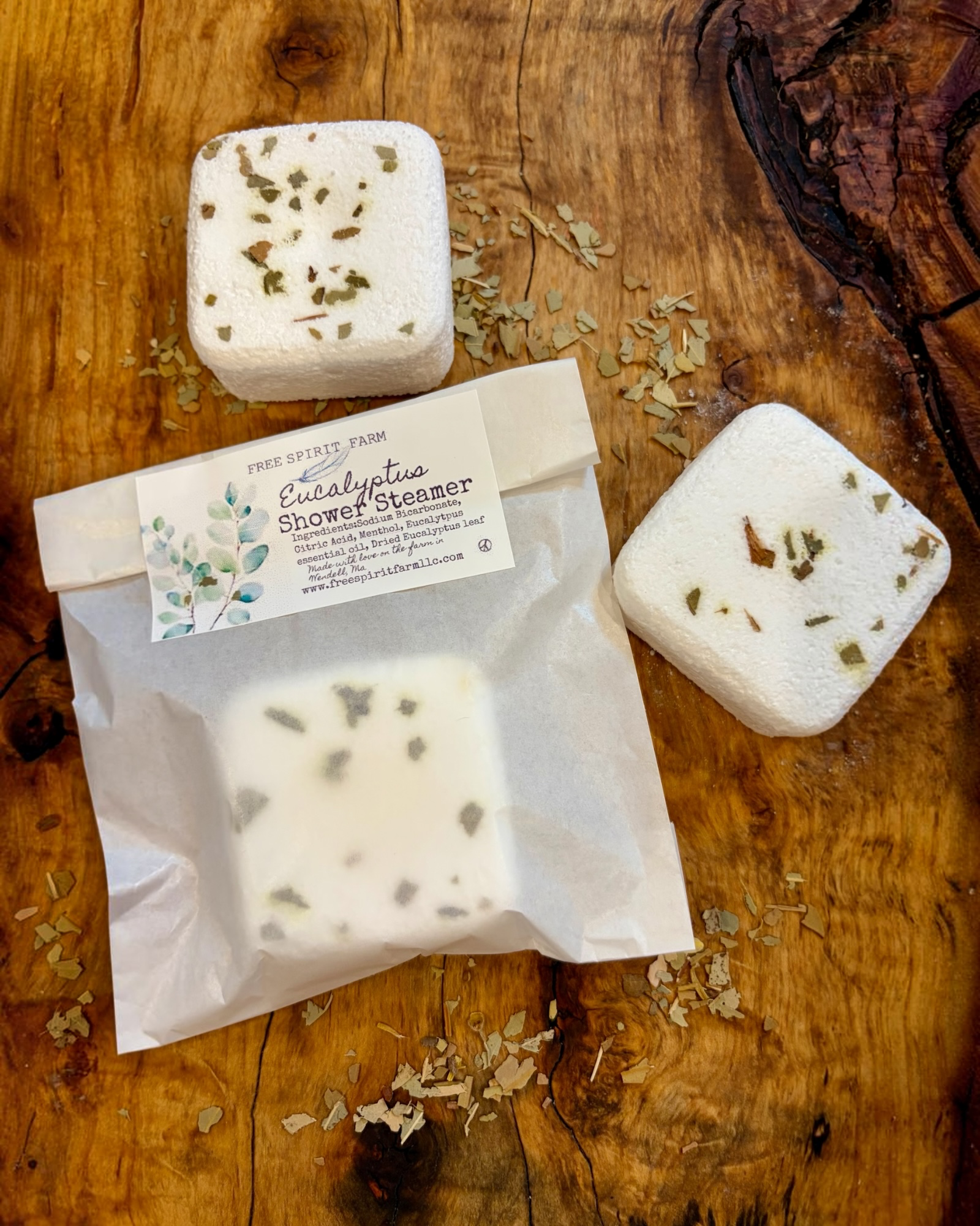 Free Spirit Farm  square white shower steamer bars with dried botanical inclusions, one shown unpackaged and one inside a clear resealable pouch with a white label reading &