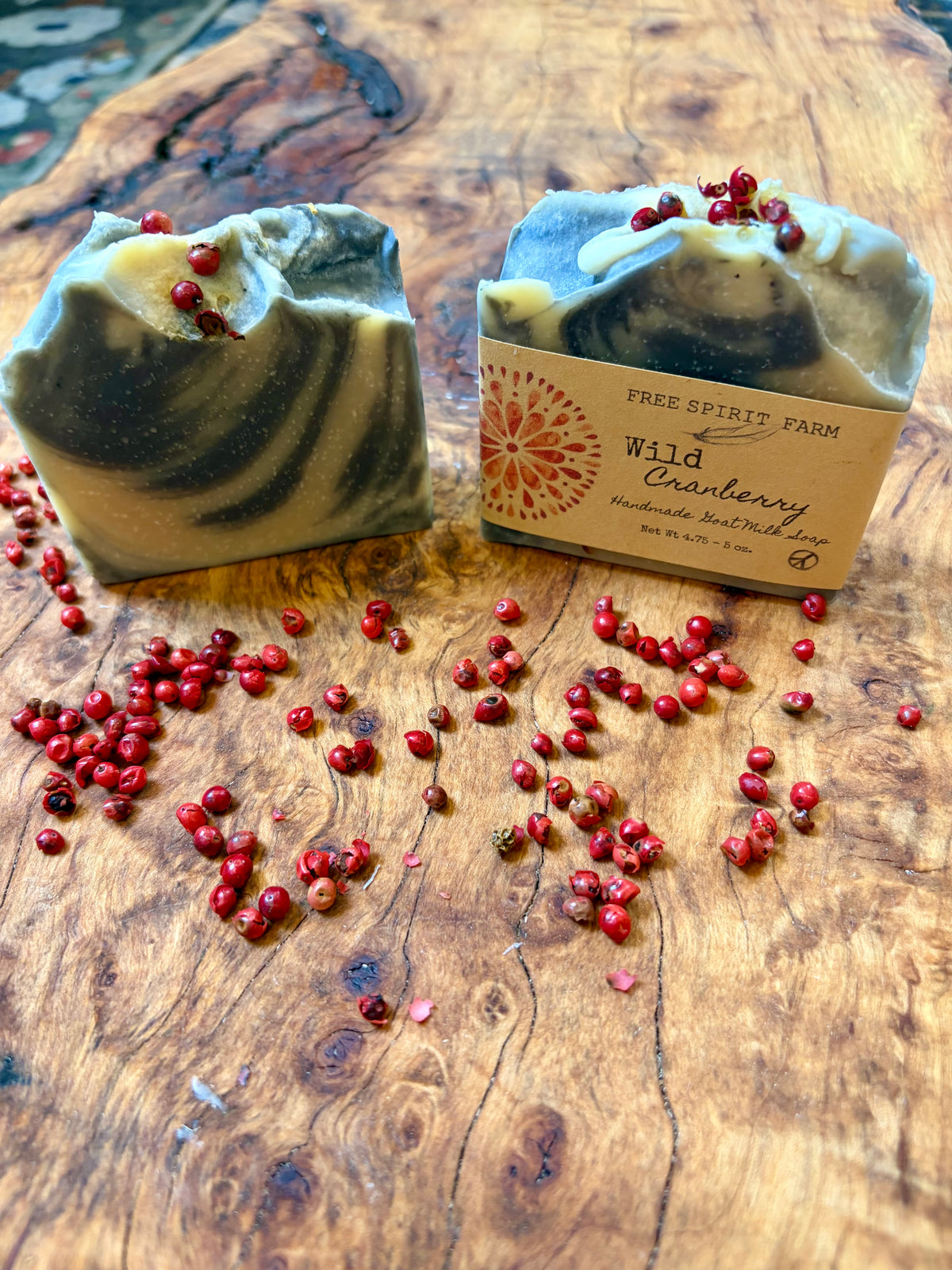 Two artisan soap bars with a dark purple and cream swirl pattern, topped with dried peppercorns, displayed on a rustic wooden surface with scattered cranberries