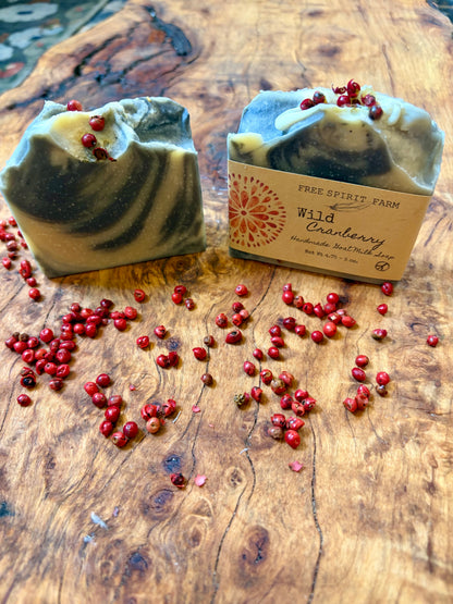Two artisan soap bars with a dark purple and cream swirl pattern, topped with dried peppercorns, displayed on a rustic wooden surface with scattered cranberries