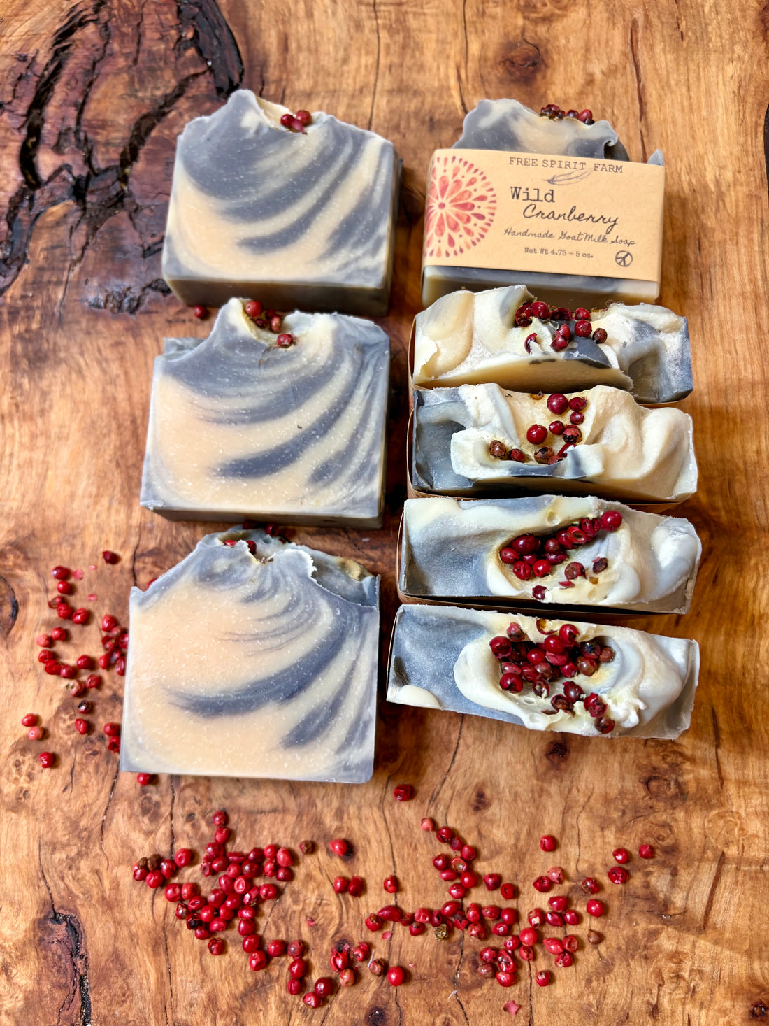  Free Spirit Farm handmade soap bars and one wrapped bar are arranged on a rustic wooden surface with scattered red cranberry seeds.