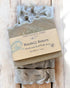 A bar of goat milk soap with exfoliating pumice on top, wrapped in a paper labeled &