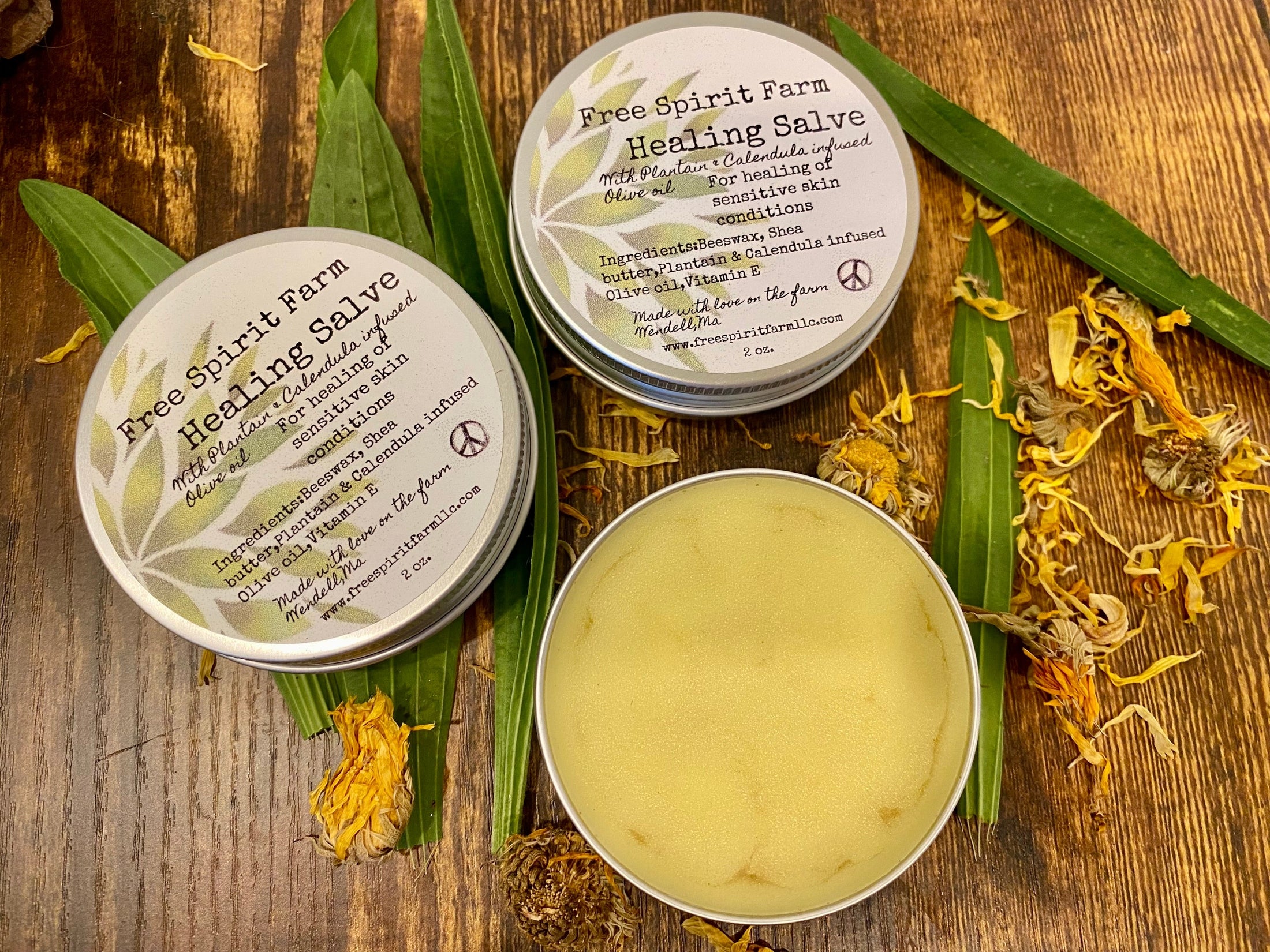 Two travel tins of Free Spirit Farm Healing Salve with Plantain &amp; Calendula, alongside a third tin that is partially melted, displaying the product. The tins are on a wooden surface with green and orange botanical elements surrounding them.