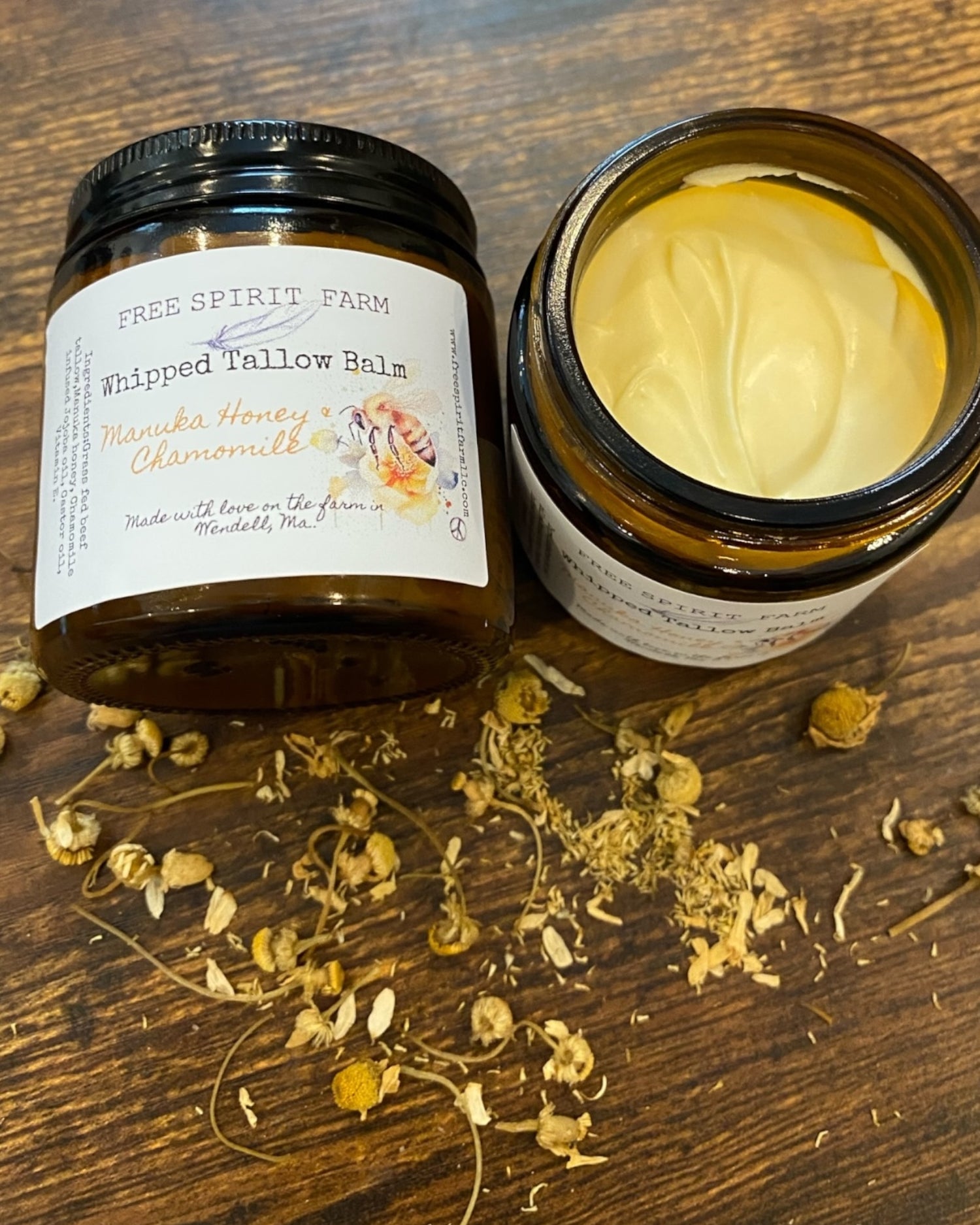 A jar of Whipped Tallow Balm with Manuka Honey &amp; Chamomile on a wooden surface, with ingredients like chamomile flowers scattered around.