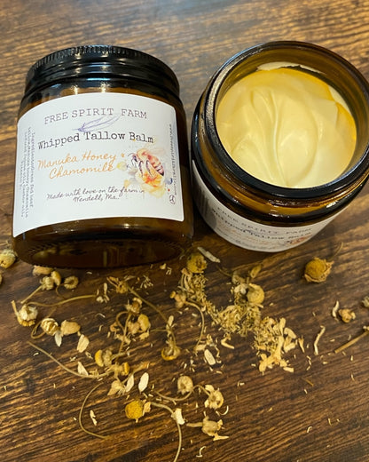 A jar of Whipped Tallow Balm with Manuka Honey &amp; Chamomile on a wooden surface, with ingredients like chamomile flowers scattered around.