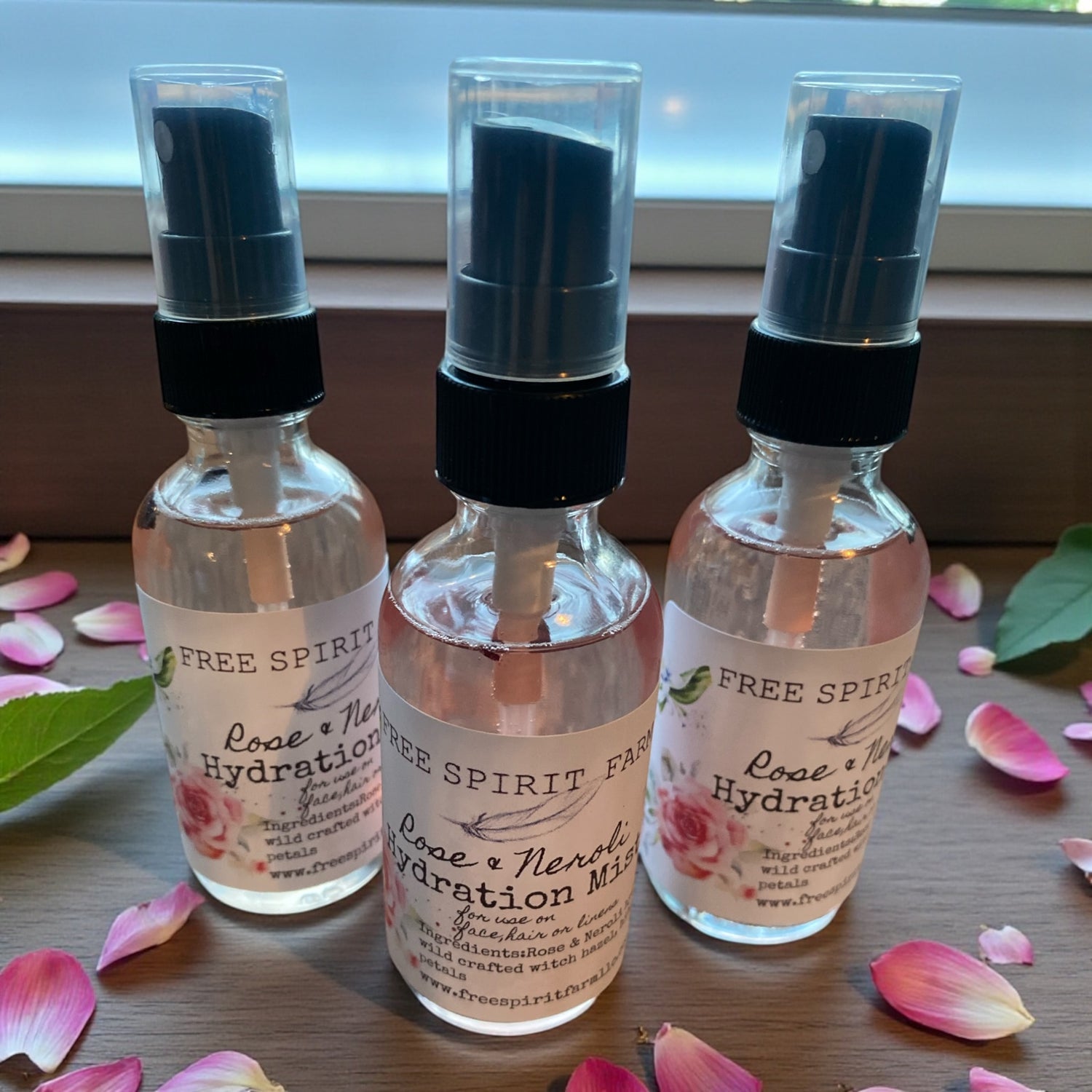 Three bottles of Free Spirit Rose &amp; Neroli Hydration Mist on a wooden surface with rose petals.