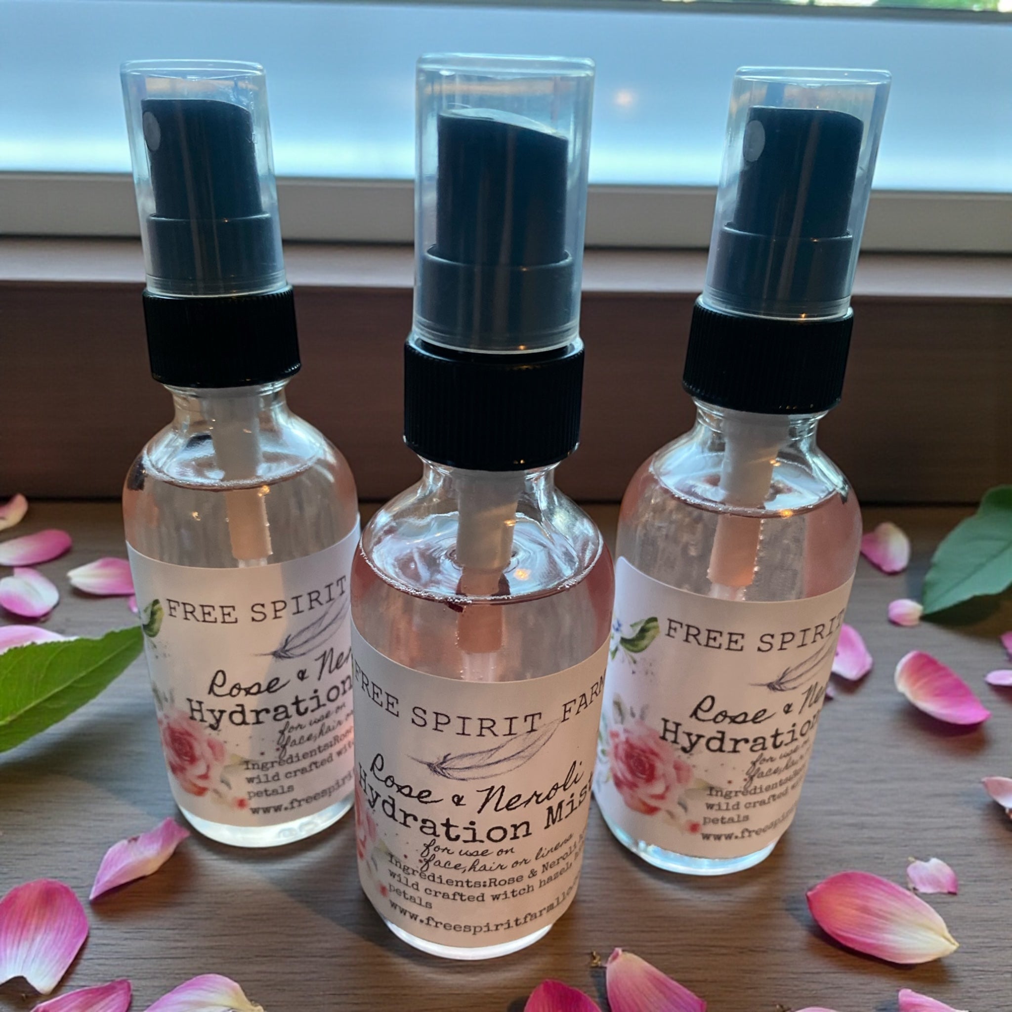 Three bottles of Free Spirit Rose &amp; Neroli Hydration Mist on a wooden surface with rose petals.
