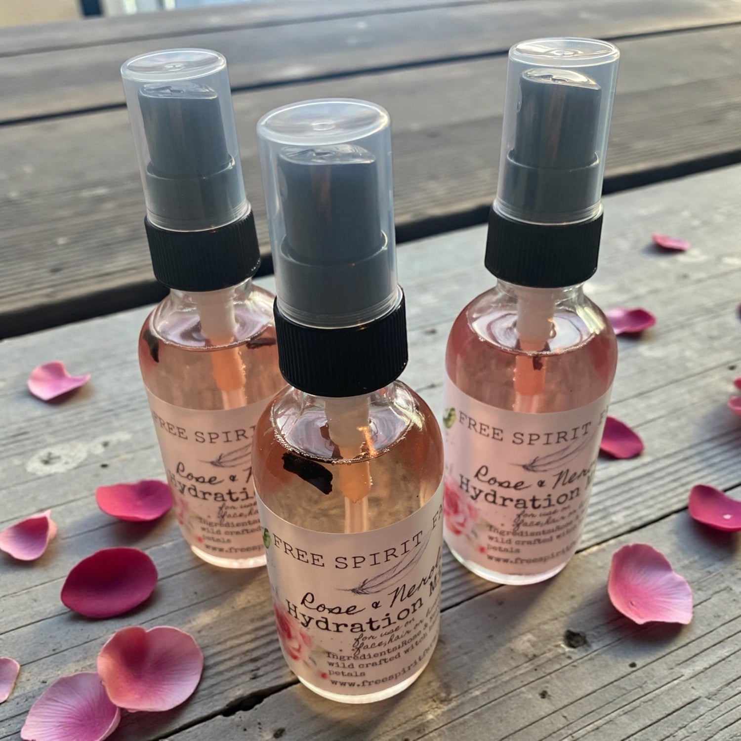 Three bottles of Free Spirit Rose &amp; Neroli Hydration spray on a wooden surface with pink petals.