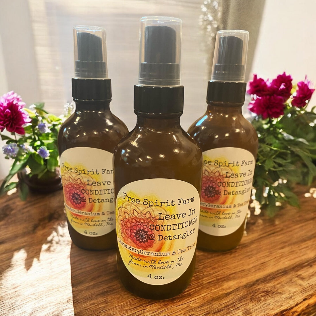 Three bottles of Free Spirit Farm Leave In Conditioner &amp; Detangler on a wooden surface with flowers in the background.