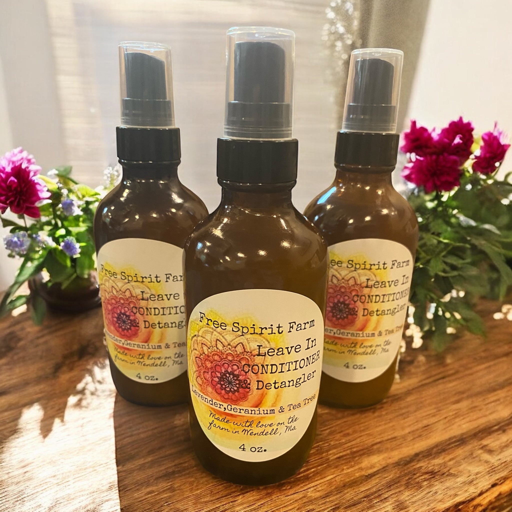 Three bottles of Free Spirit Farm Leave In Conditioner &amp; Detangler on a wooden surface with flowers in the background.