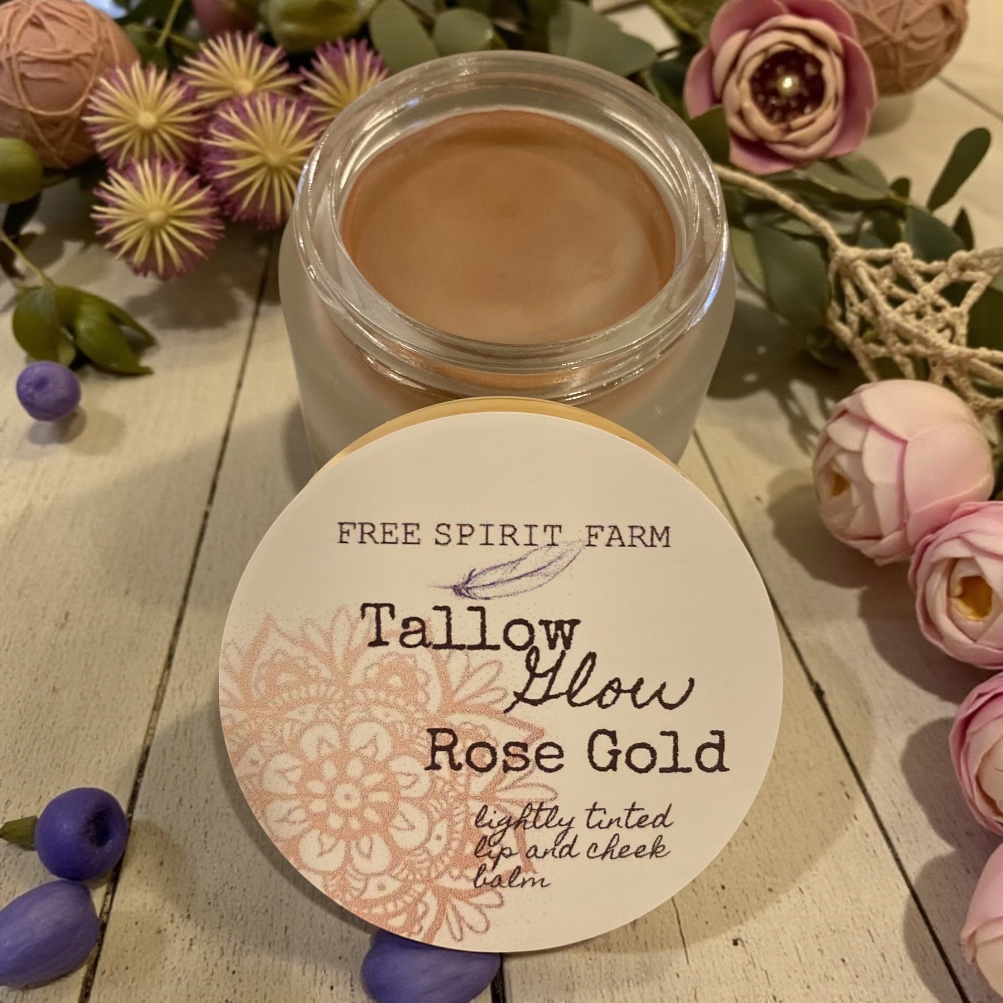 Tallow Glow Rose Gold balm from Free Spirit Farm on a wooden surface with flowers and berries.