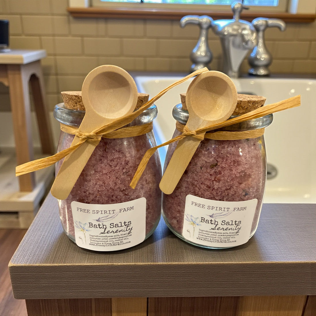 Two jars of bath salts with wooden spoons on a bathroom counter.