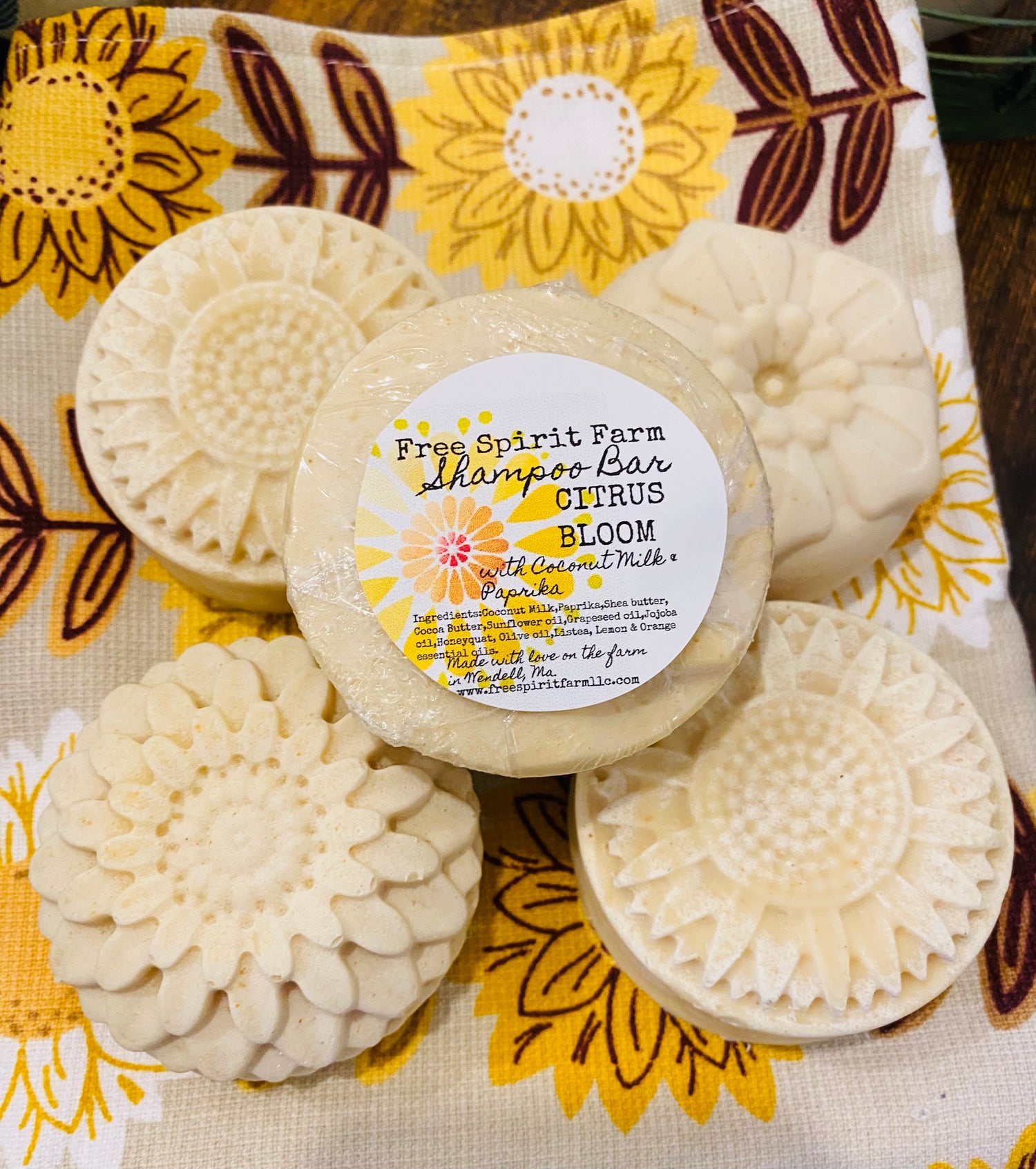 Three shampoo bars wrapped in biodegradable shrink wrap, with a label in the center displaying the product name and ingredients, placed on a fabric with a floral pattern.