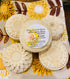 Three shampoo bars wrapped in biodegradable shrink wrap, with a label in the center displaying the product name and ingredients, placed on a fabric with a floral pattern.