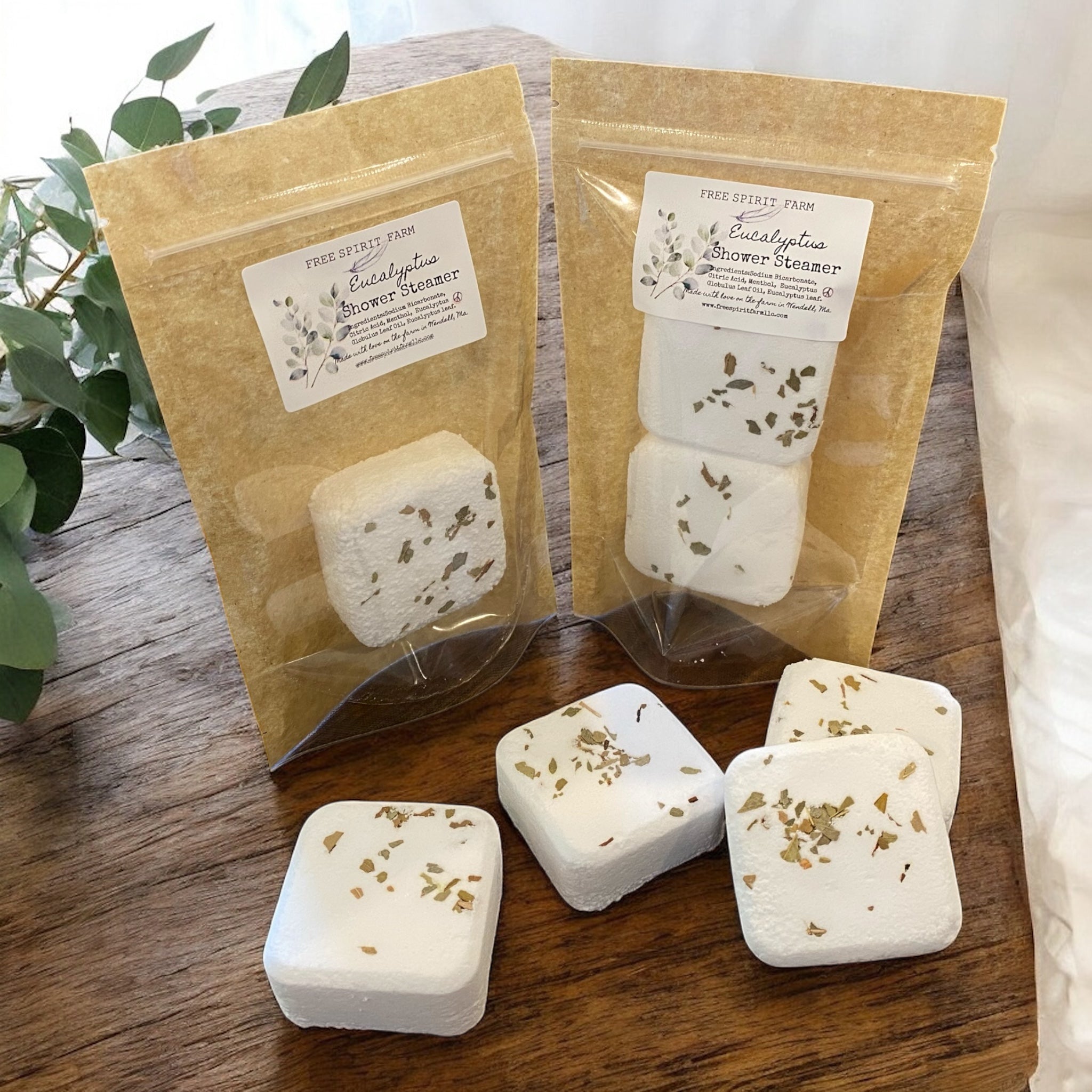 Two packages of Eucalyptus Shower Steamers on a wooden surface with eucalyptus leaves in the background.
