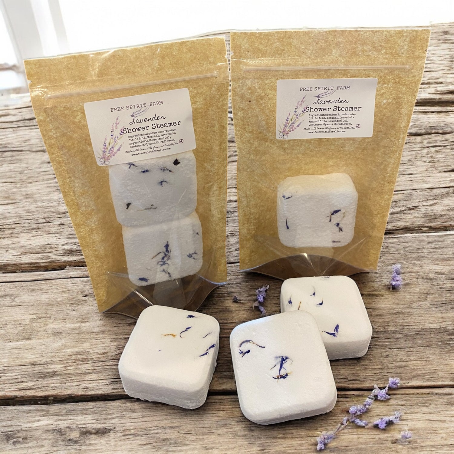 Two packages of lavender shower steamers on a wooden surface with scattered lavender flowers.