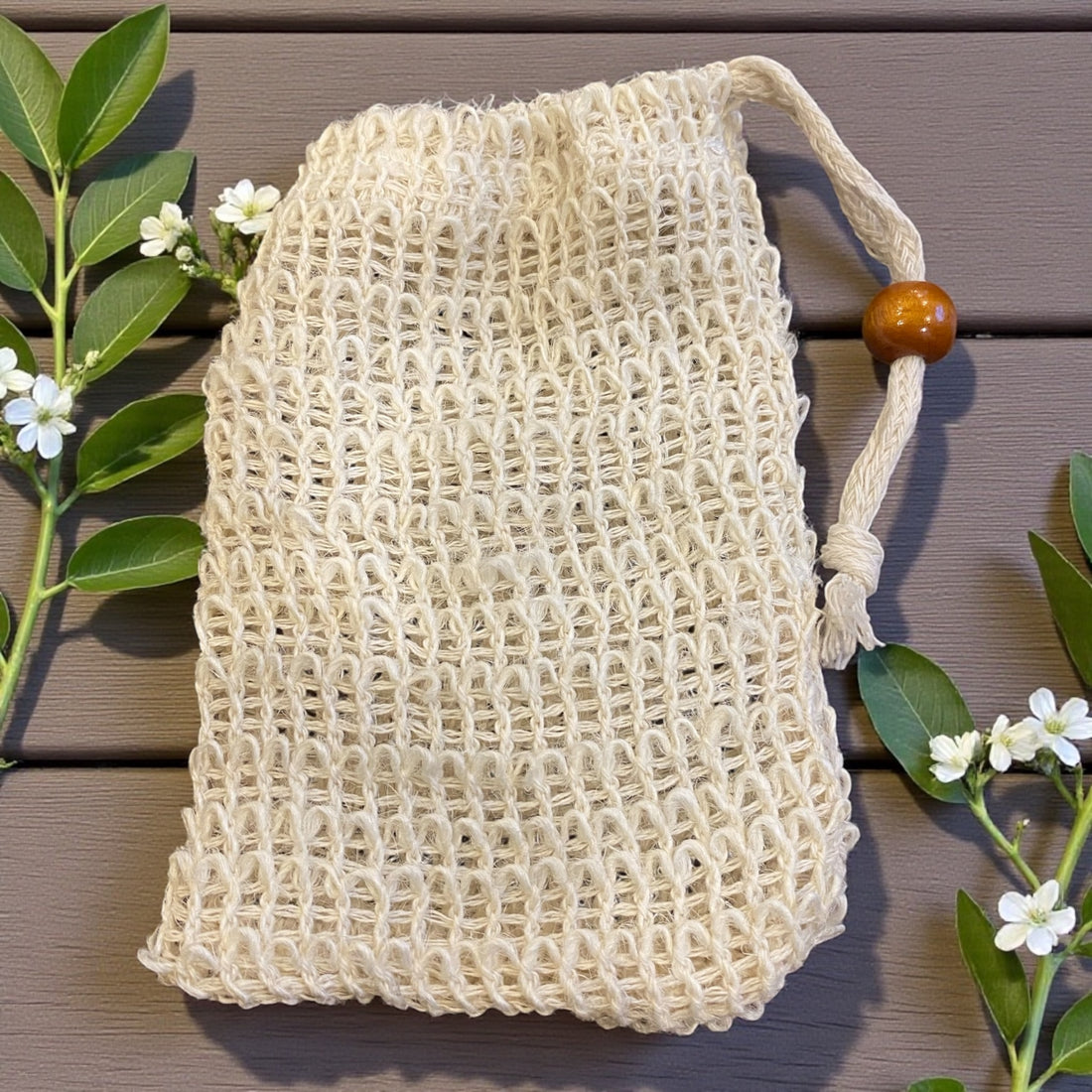 Beige mesh bag with a wooden handle on a wooden surface with green leaves and flowers.