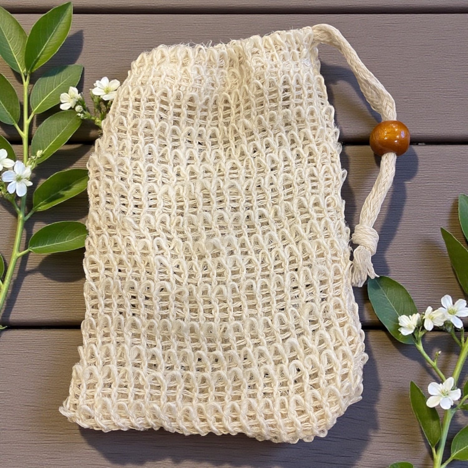 Beige mesh bag with a wooden handle on a wooden surface with green leaves and flowers.
