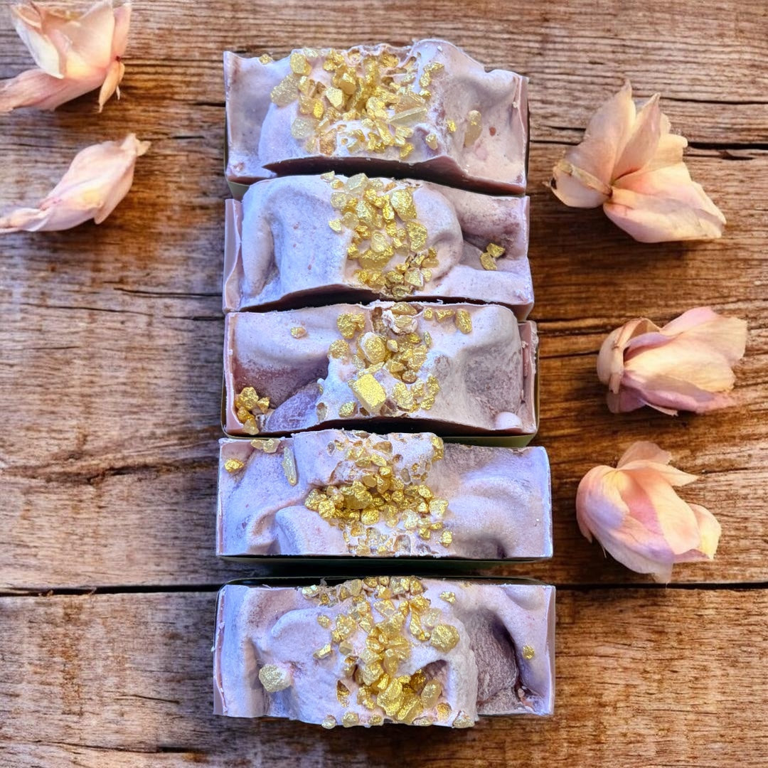 Five soap bars with gold flakes on a wooden surface surrounded by pink flowers.