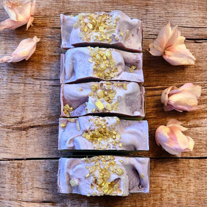 Five soap bars with gold flakes on a wooden surface surrounded by pink flowers.
