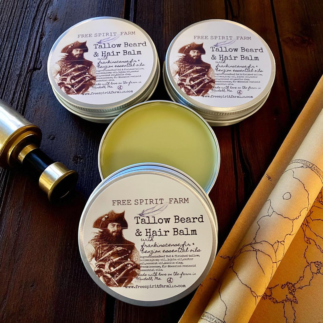 Three tins of Free Spirit Farm Tallow Beard &amp; Hair Balm on a wooden surface with a map and bottle.