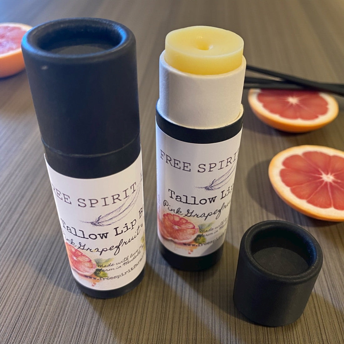Two tallow lip balm tubes with black caps on a wooden surface with grapefruit slices.