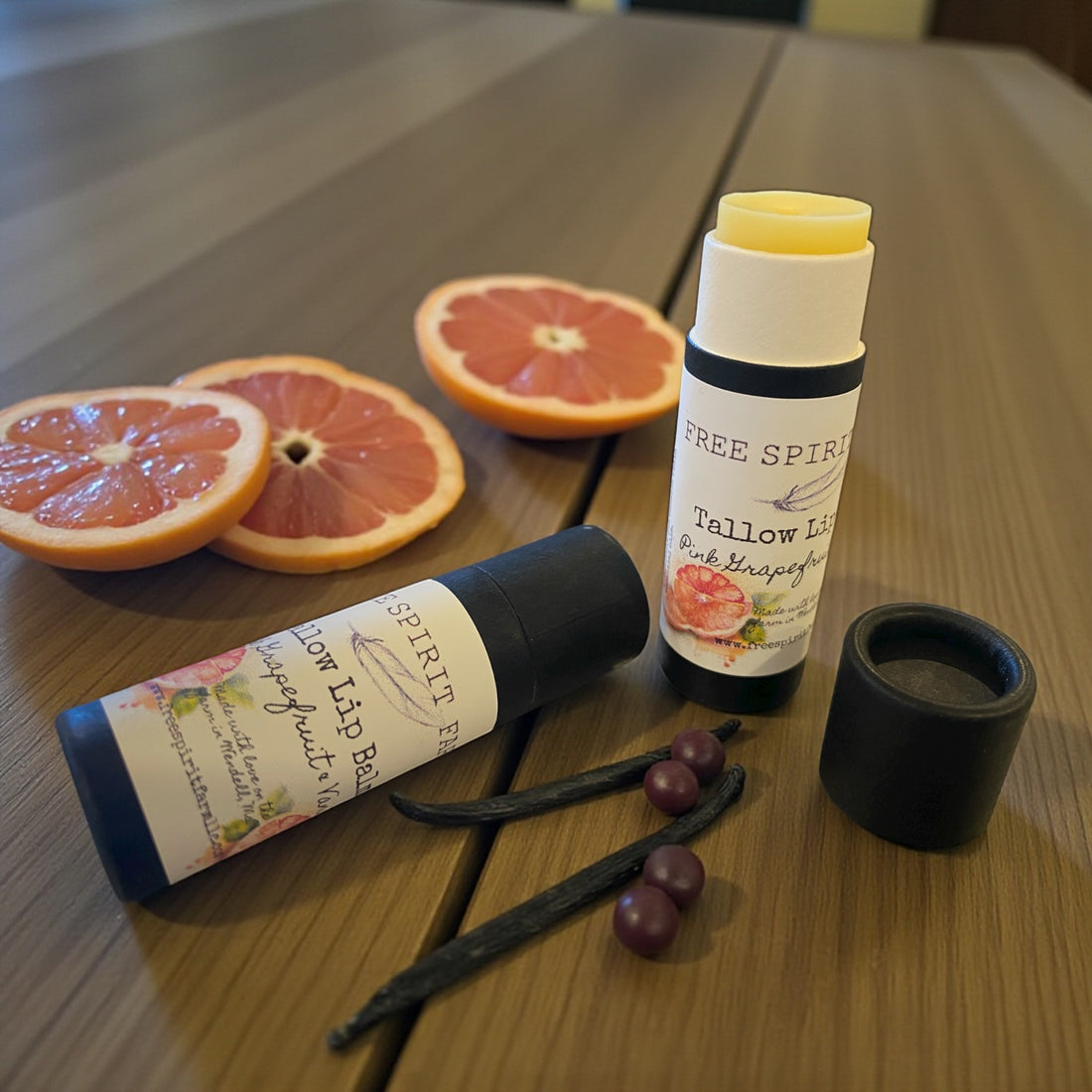 Two lip balm tubes on a wooden surface with sliced grapefruits and vanilla beans.