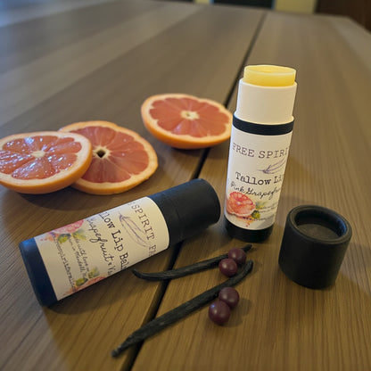Two lip balm tubes on a wooden surface with sliced grapefruits and vanilla beans.