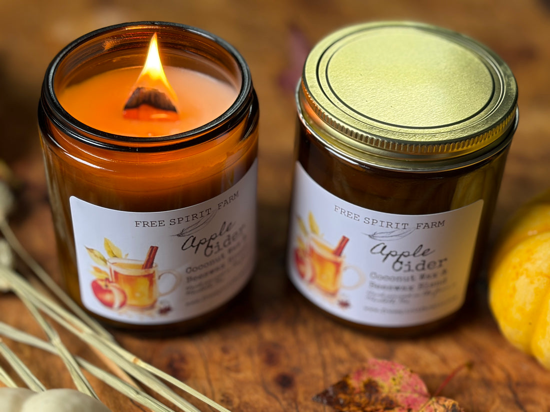 wo amber glass jars of scented candles from Free Spirit Farm, one lit with a visible flame and one sealed with a gold lid, both labeled Apple Cider, set on a rustic wooden surface with dried grass and autumn leaves.