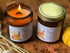 wo amber glass jars of scented candles from Free Spirit Farm, one lit with a visible flame and one sealed with a gold lid, both labeled Apple Cider, set on a rustic wooden surface with dried grass and autumn leaves.
