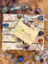 A set of handmade soap bars with embedded gemstones, arranged on a wooden surface with loose crystals around them. A paper label on one bar reads &