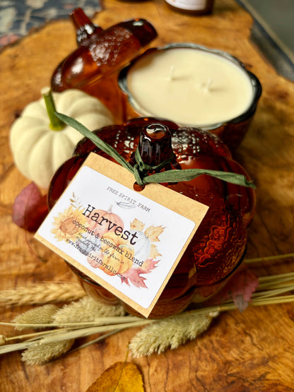 A scented candle in a decorative amber glass pumpkin container sits on a rustic wooden surface, styled with dried grasses, a small white pumpkin, and a second candle in a matching amber holder. A paper tag reads &