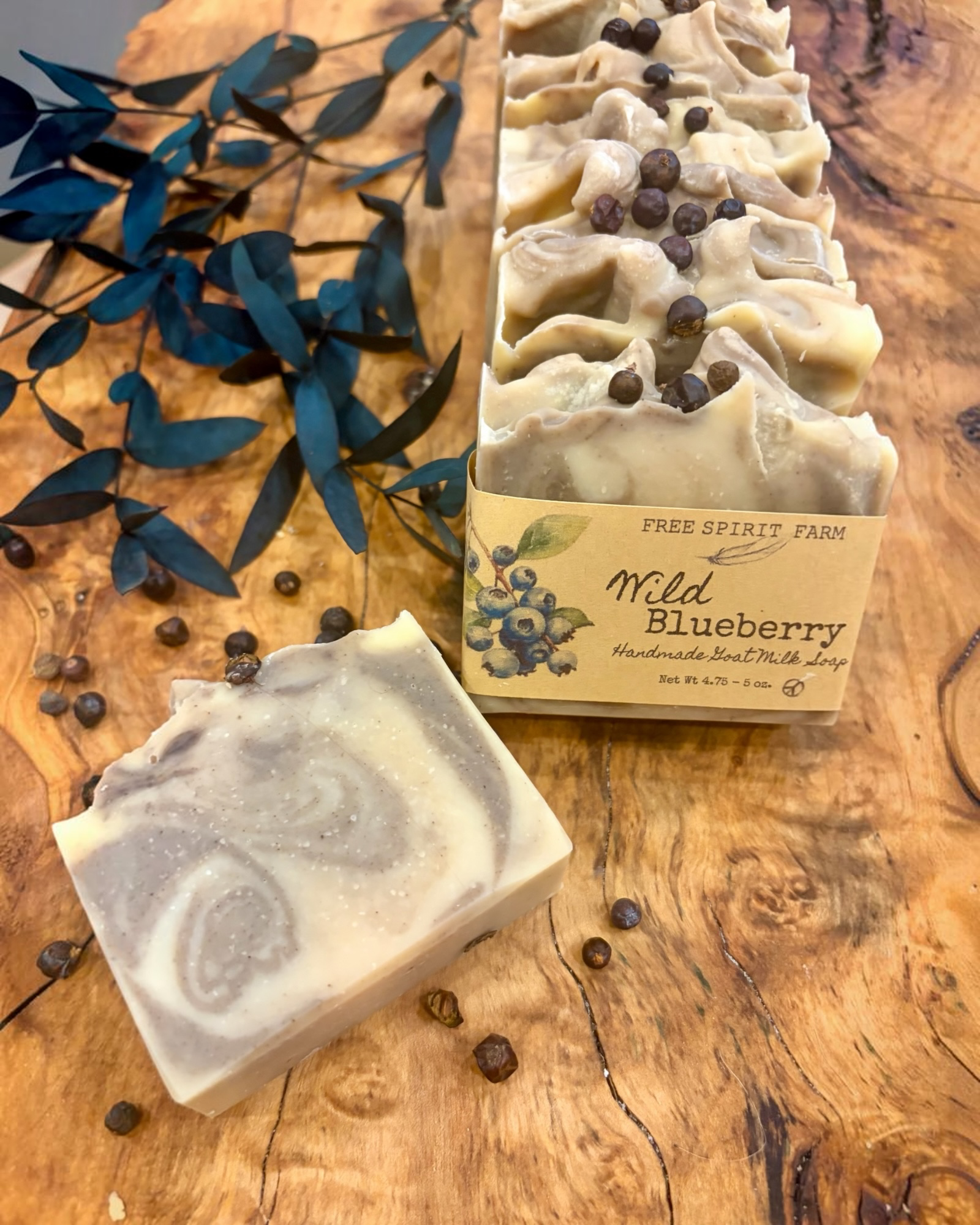 A handmade soap bar and a matching bar in a paper sleeve are displayed on a rustic wooden surface with scattered blueberries and blue-green leaves. The soap has a swirled cream and blue design, and the packaged soap is topped with whole juniper berries.